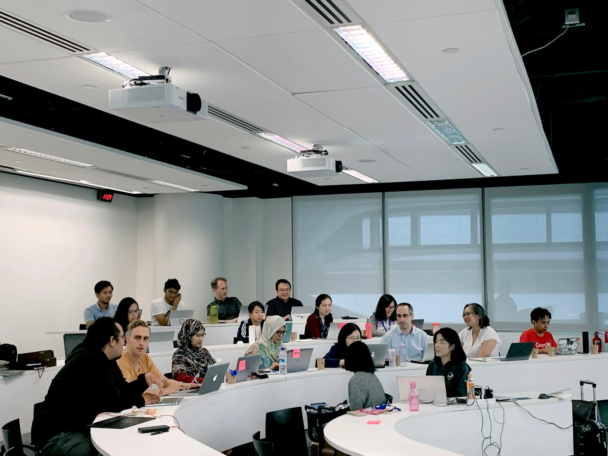 Students with laptops at curved tables in a bright seminar room, listening to someone speak
