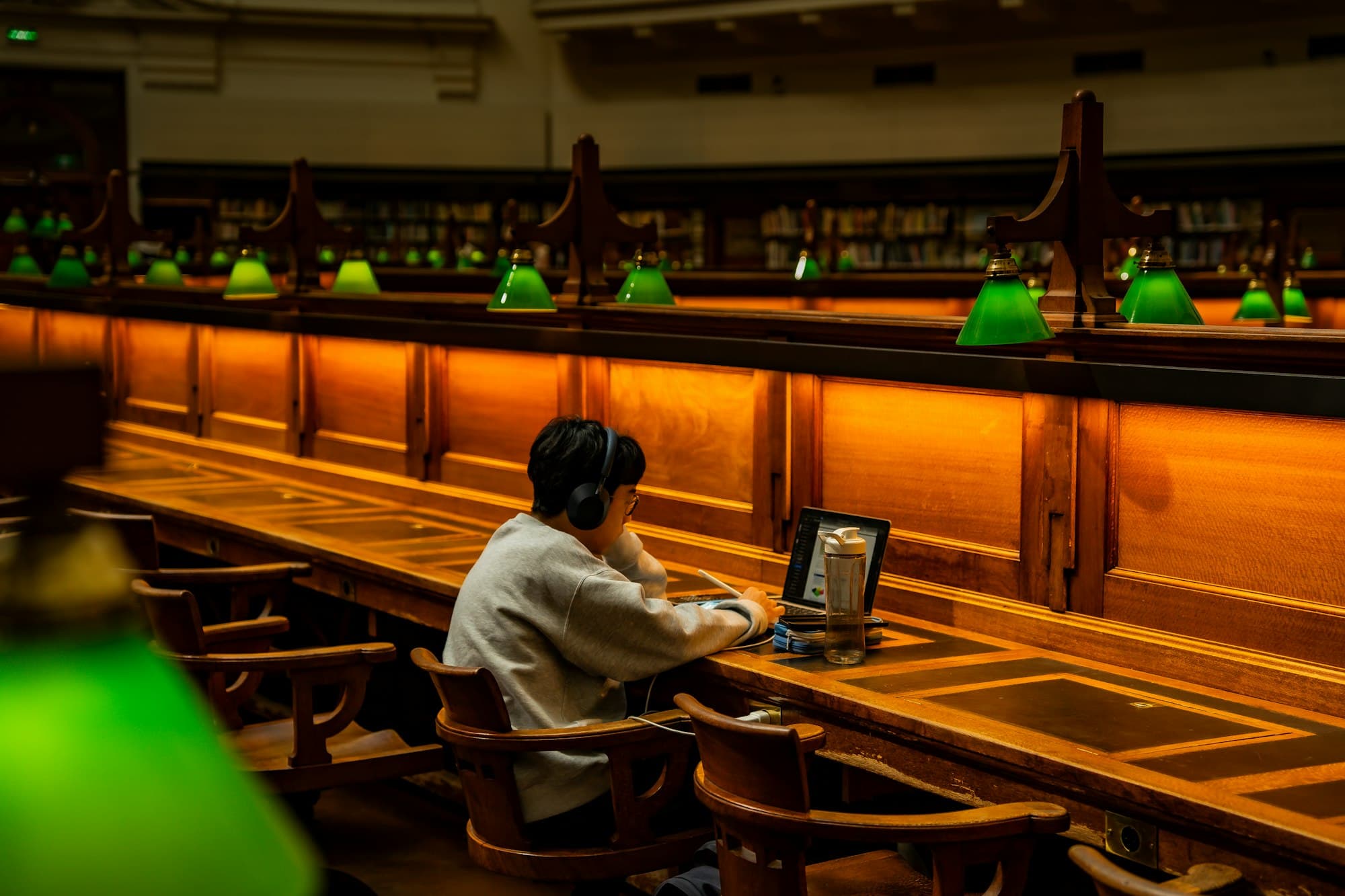 A student working on a laptop at a long wooden library desk under green reading lamps