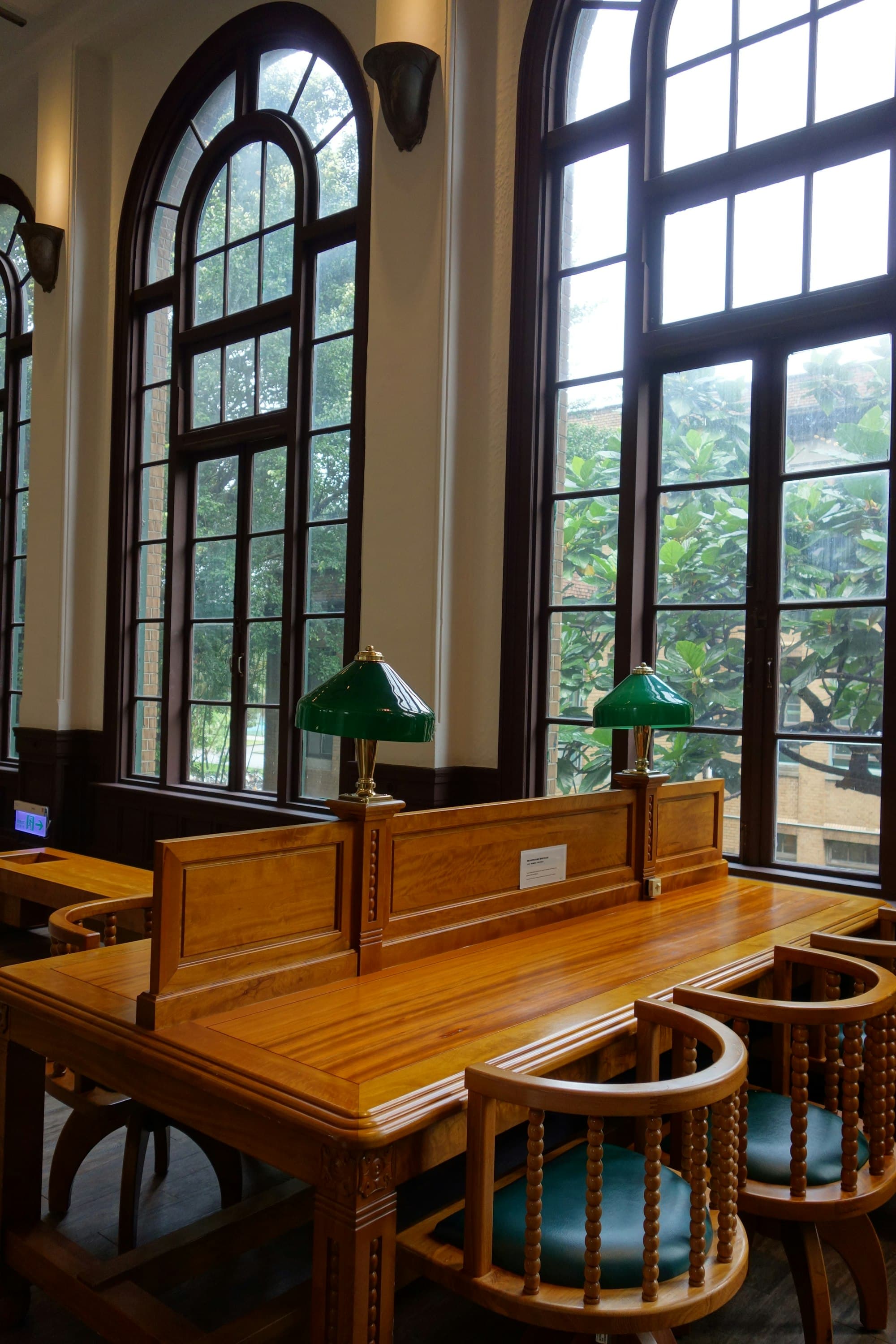 An empty reading room with tall windows, wooden desks and green lamps